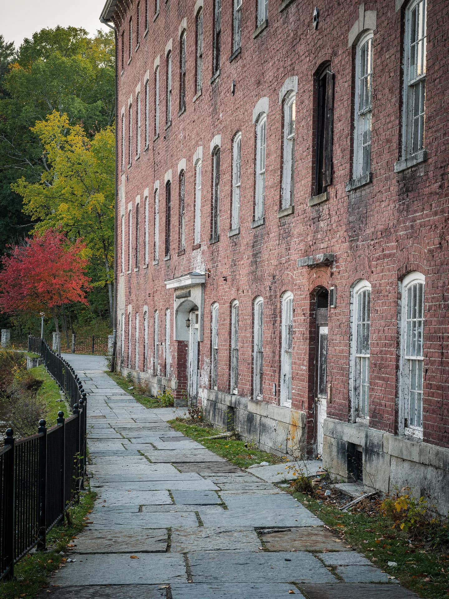 A flagstone walkway with black iron railings runs alongside a long red-brick mill wall lined with rows of tall windows.