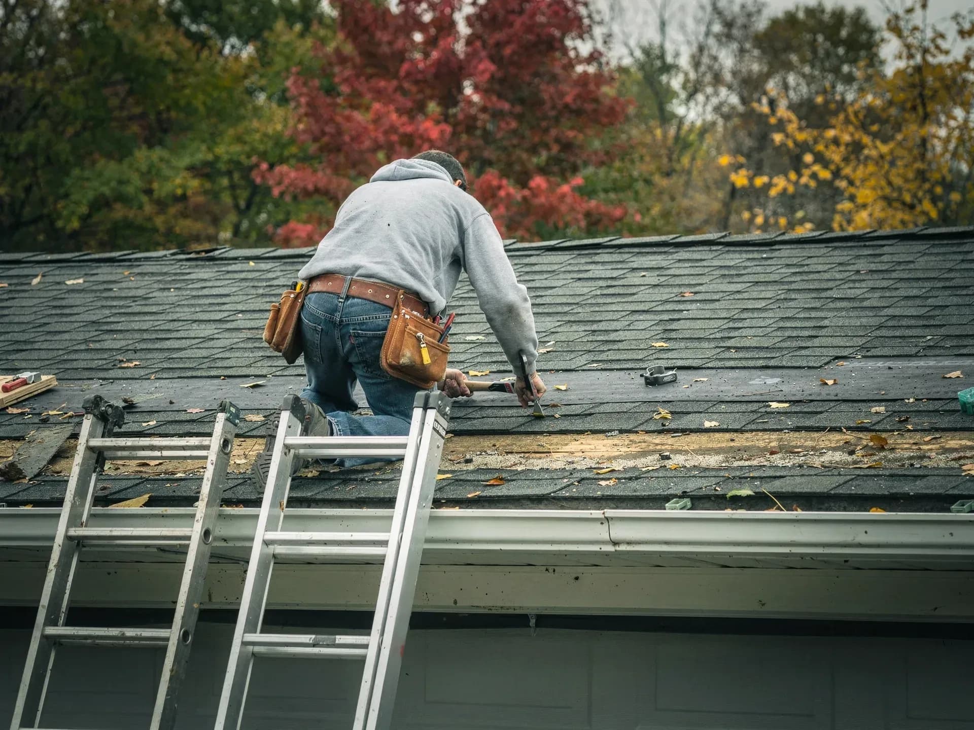 A roofer in a grey hooded sweatshirt and tool belt kneels on a garage roof nailing down dark shingles, ladder rails showing at the eave.