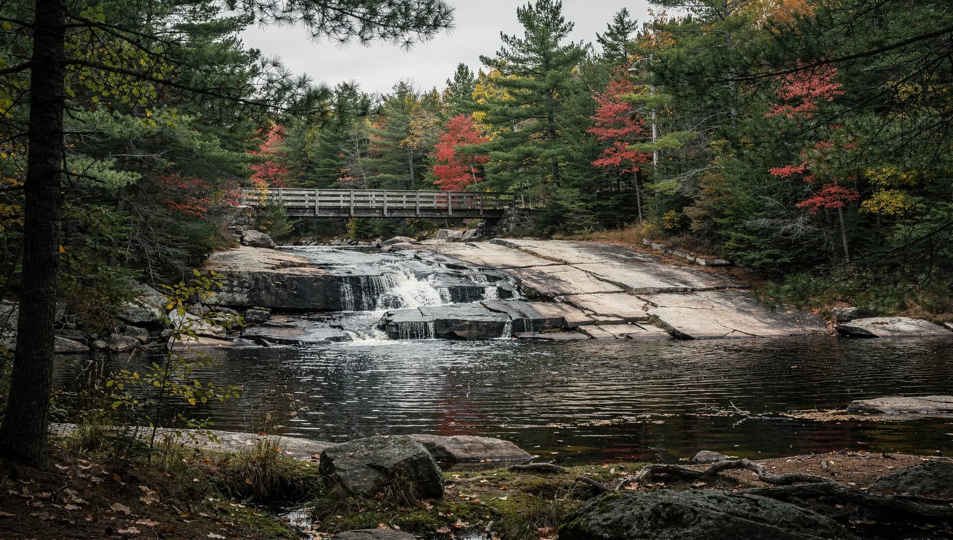 A river slides over granite ledges into a dark pool in pine woods, a wooden footbridge crossing upstream.