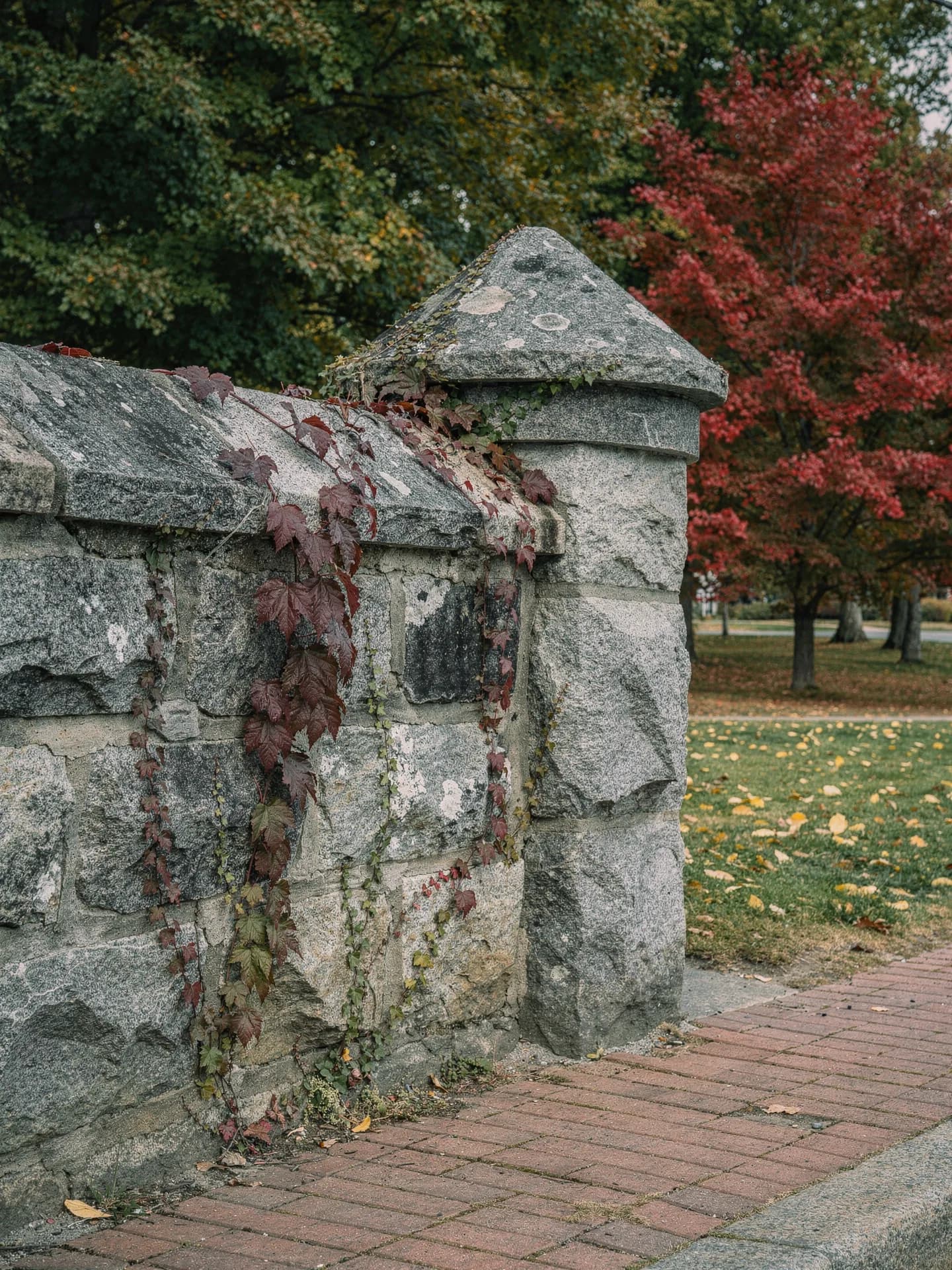 A rough granite wall ends in a small round turret with a conical cap, red ivy trailing over the stone above a brick sidewalk.