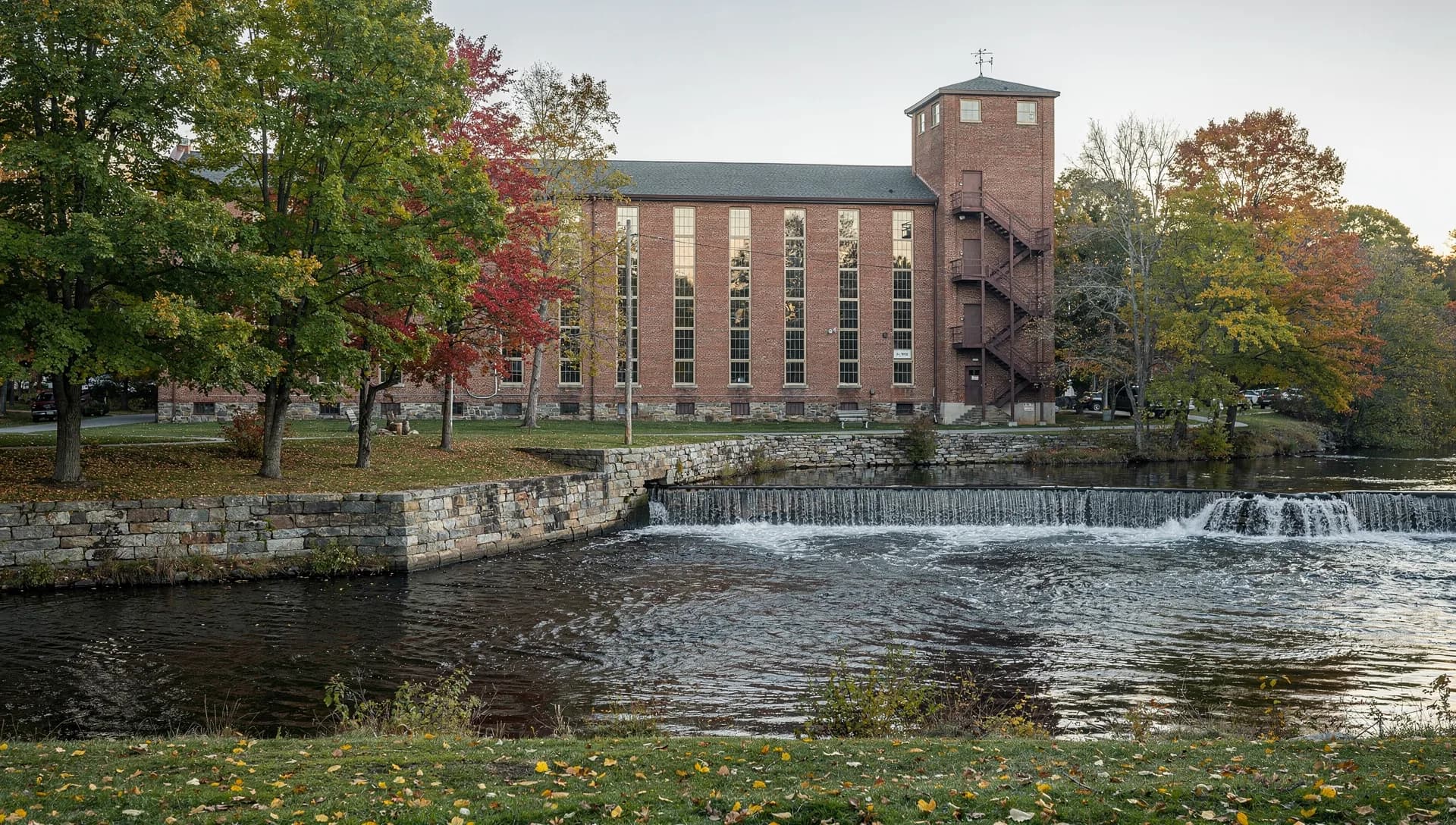 A brick mill with a stair tower stands beside a low stone dam and a granite wall.