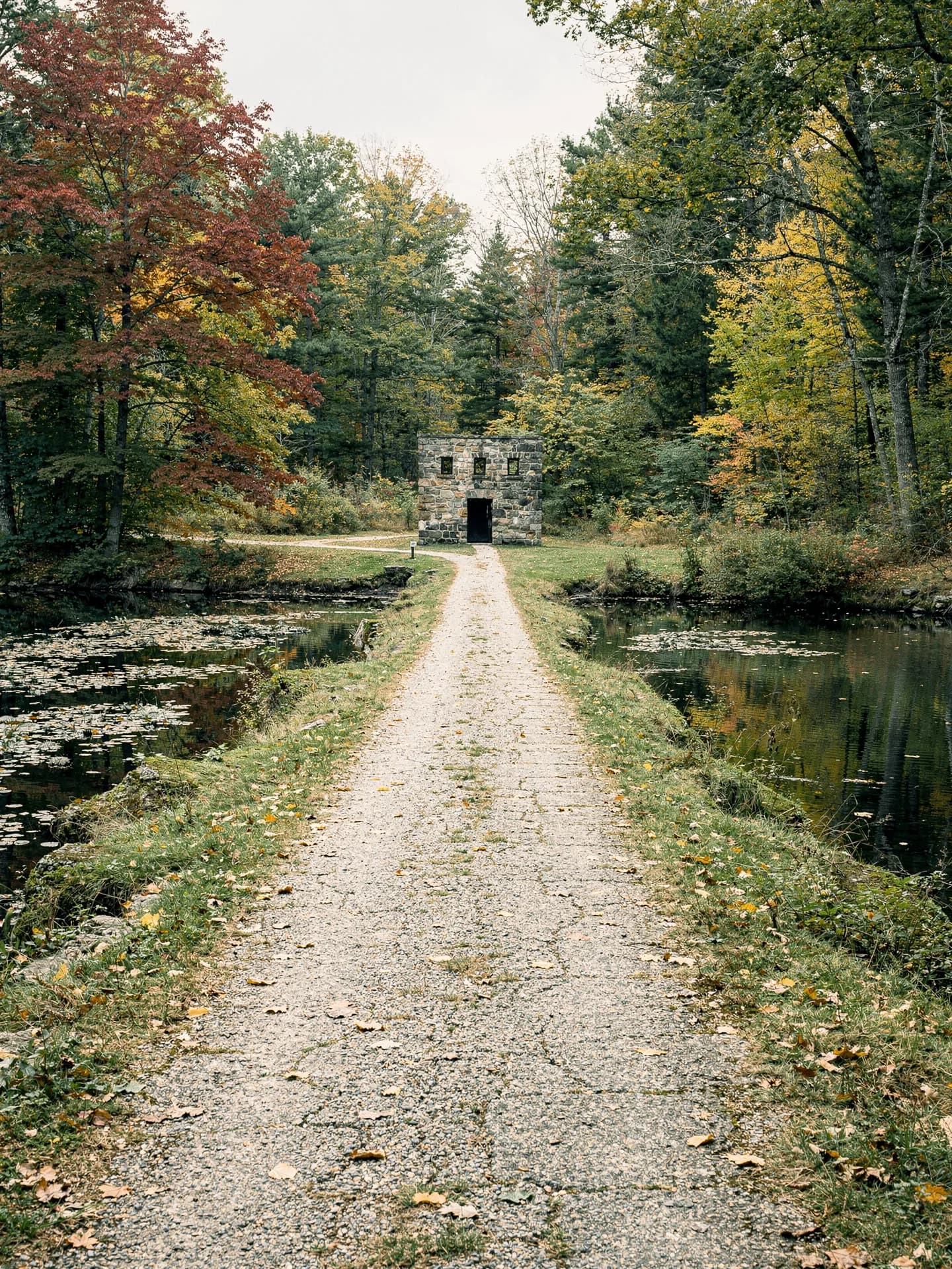 A straight gravel towpath runs between still water on both sides toward a small stone gatehouse in autumn woods.