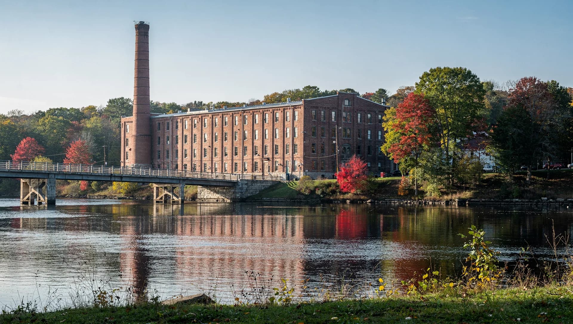 A long brick mill and a tall round chimney line a river, their reflections lying on still water beside a low bridge.