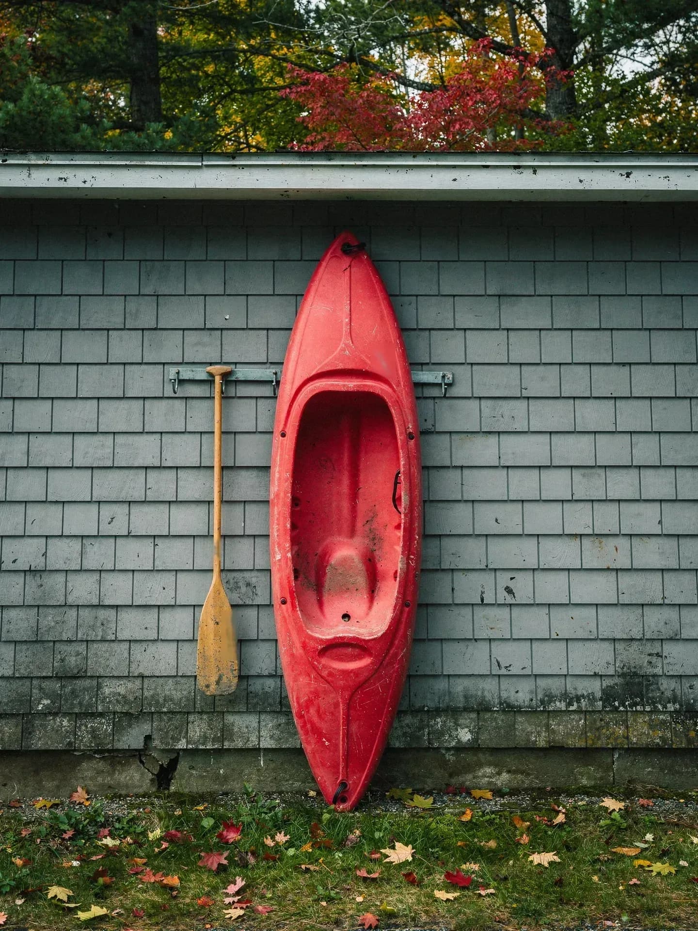 A faded red kayak leans upright against the grey shingled wall of a garage, a wooden paddle hanging on a hook beside it.