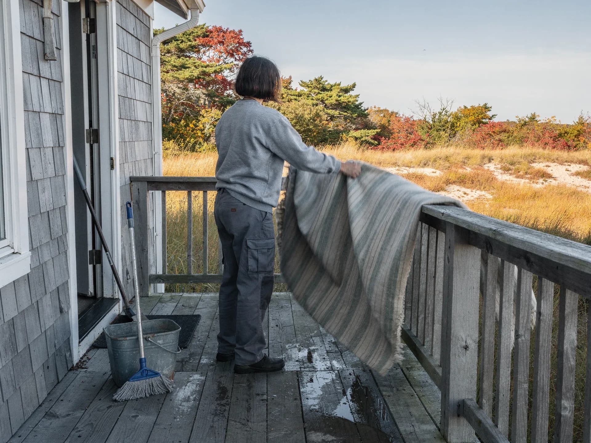 A cleaner in a grey sweatshirt shakes a striped rug over the deck rail of a shingled cottage, a mop and bucket by the door and dune grass beyond.