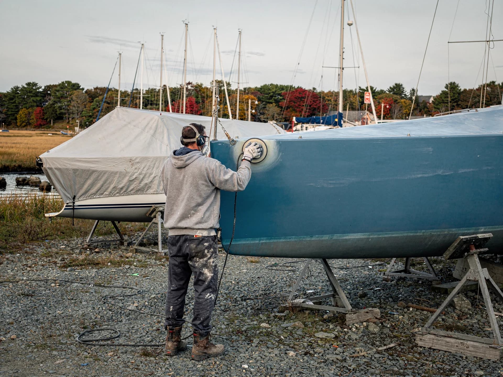 A boatyard worker in a hooded sweatshirt, ear defenders and a mask sands the bow of a blue sailboat on stands, masts and a covered boat behind.