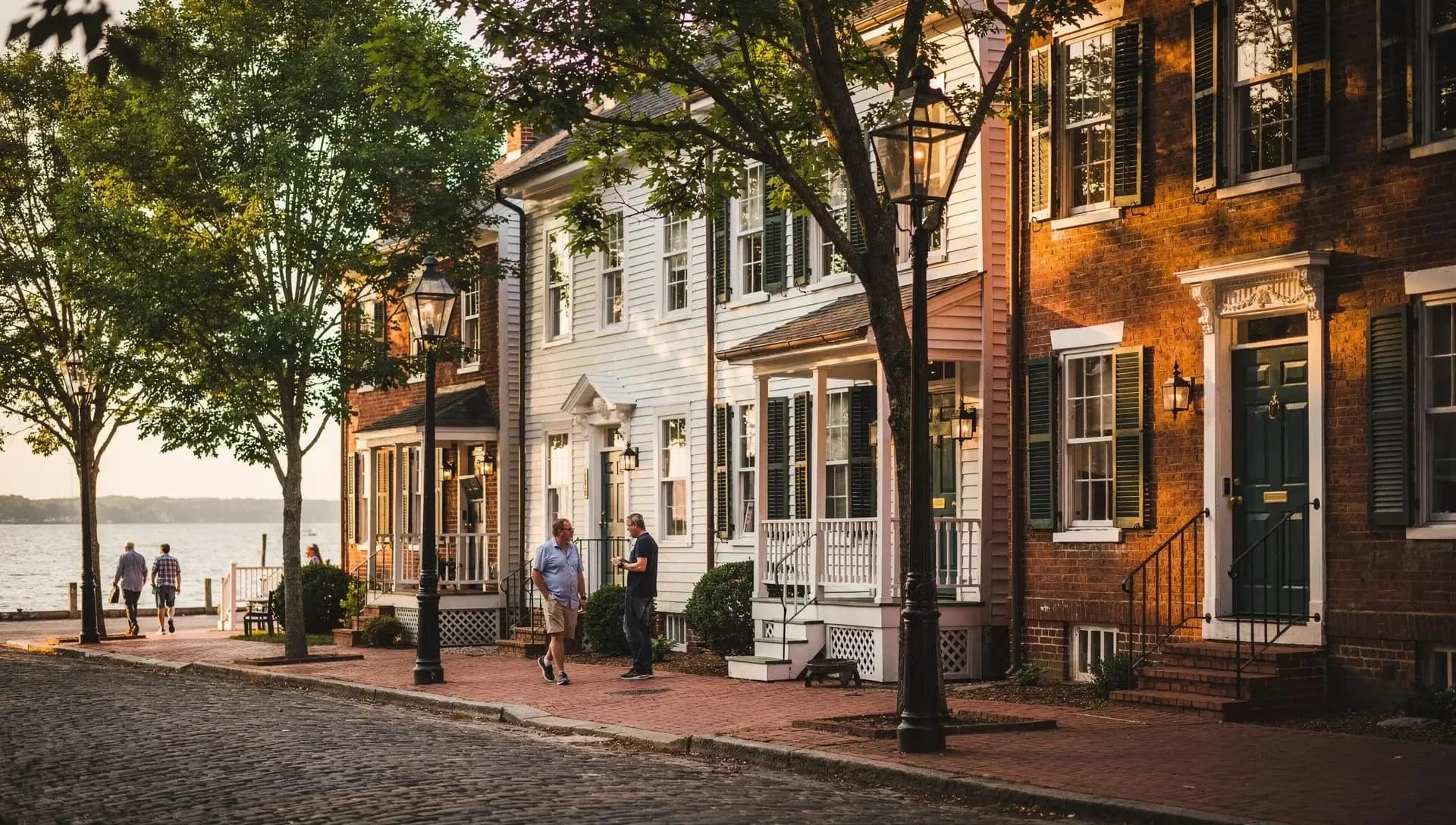 A cobblestone street of brick and white clapboard townhouses with black lamp posts, two men talking on the brick sidewalk and open water at the end of the street.