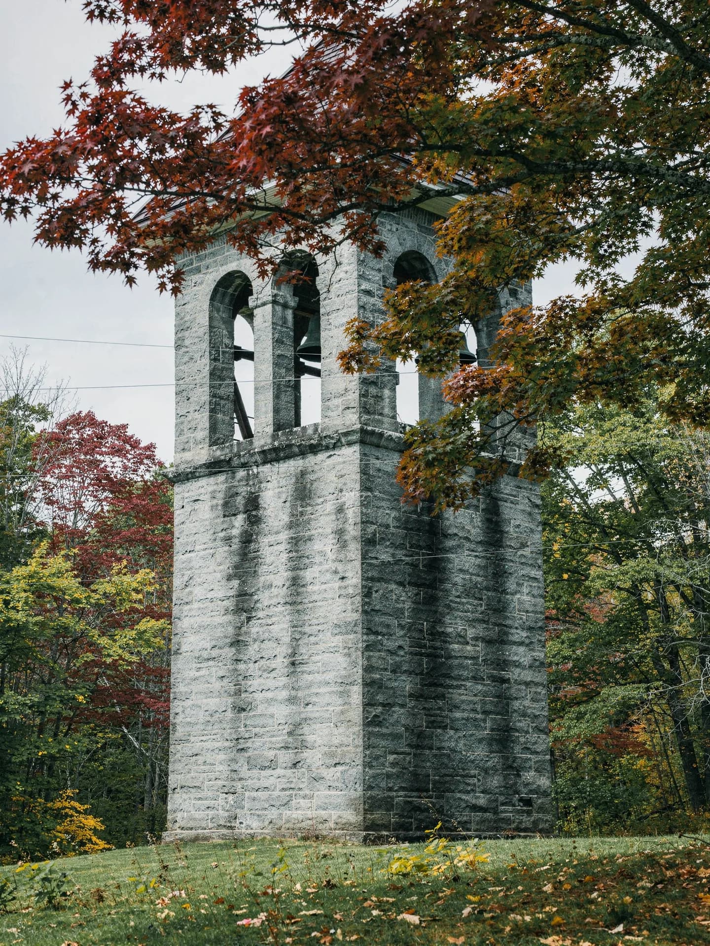 A square granite bell tower with an open arched belfry rises behind a red maple branch.