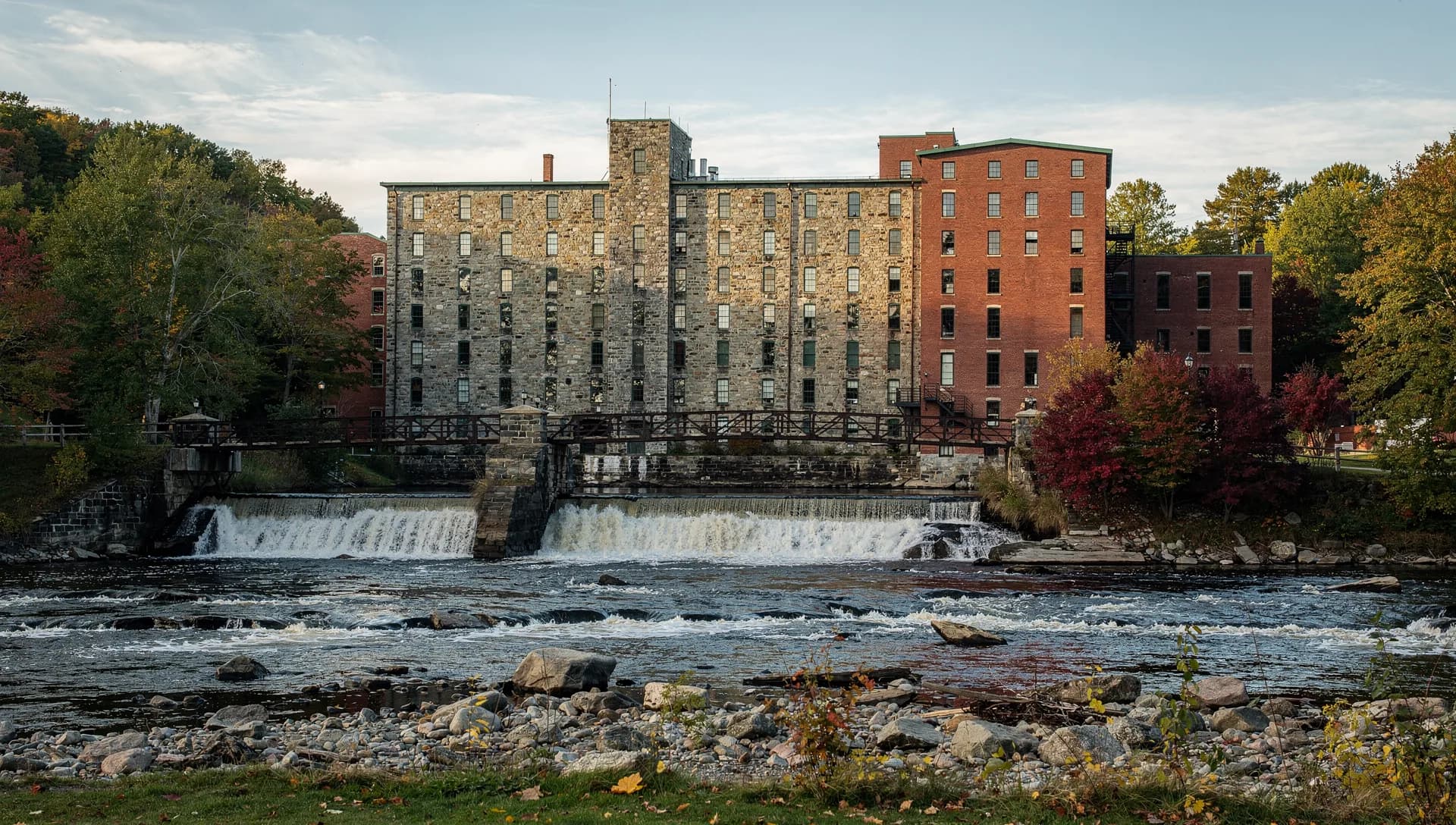 Tall stone and brick mills rise above a broad falls, an iron footbridge crossing at the dam.