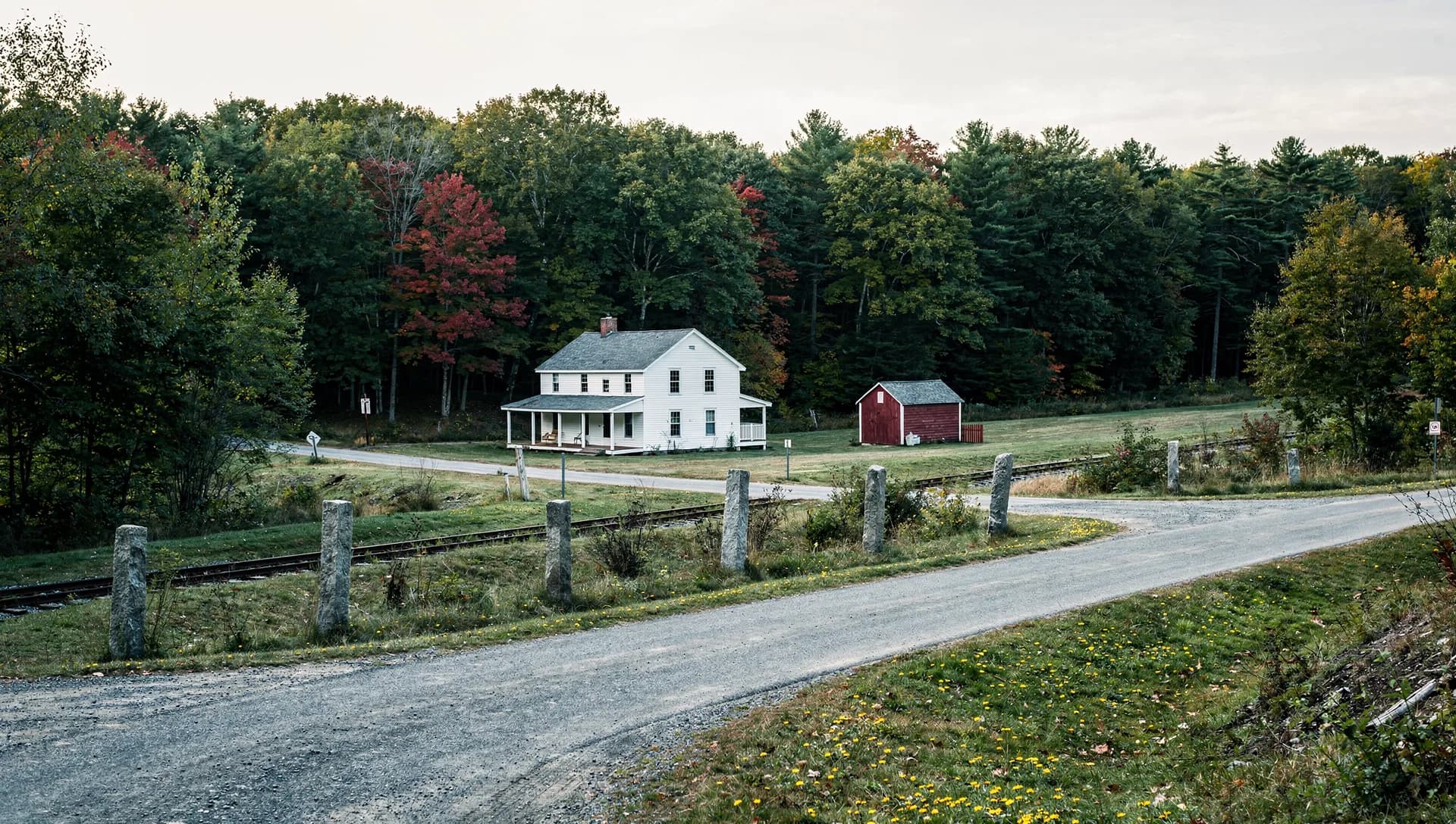 A white farmhouse with a long porch and a red shed stand at a country crossroads, granite posts marking an old rail line.