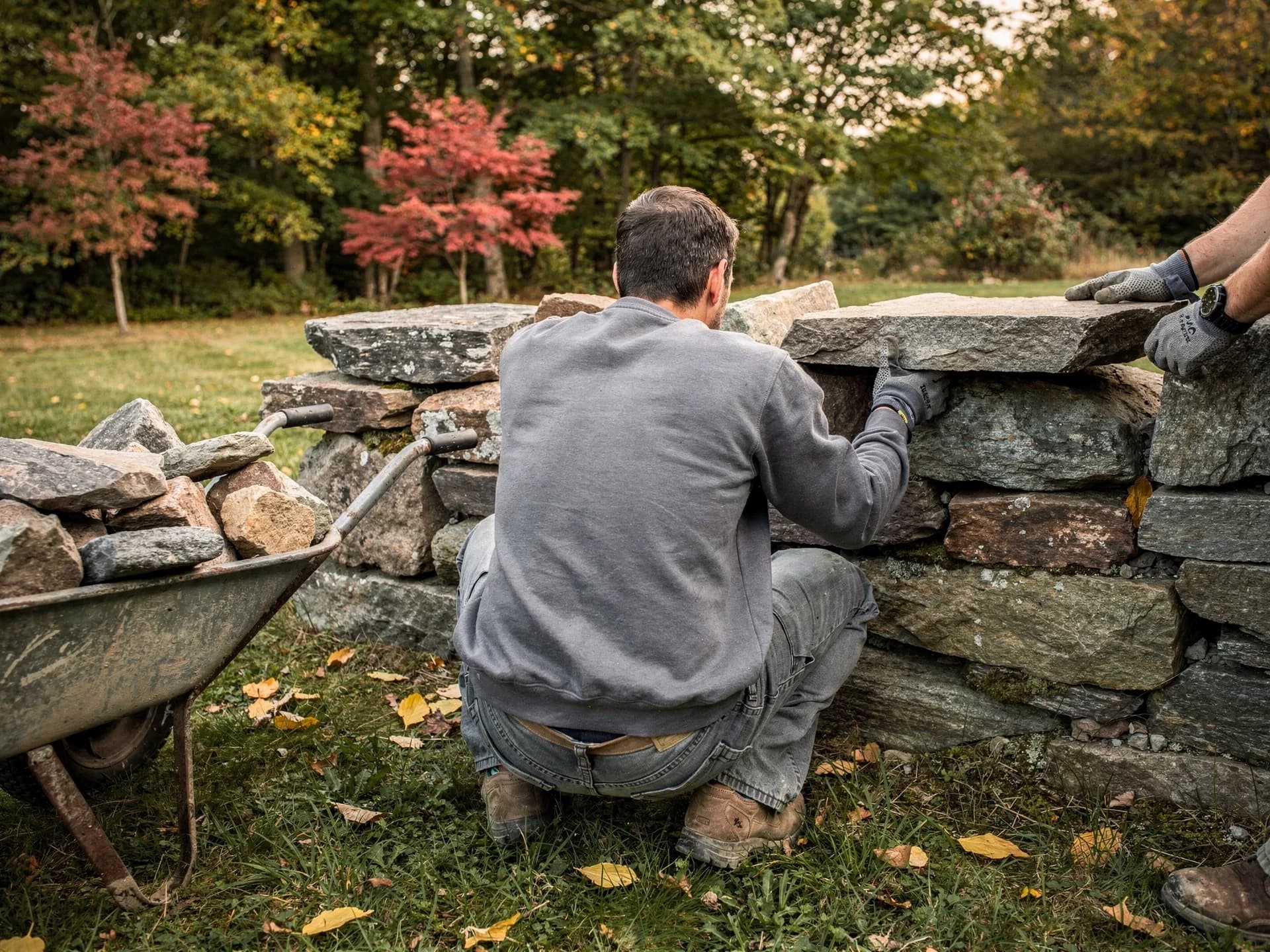A mason in a grey sweatshirt crouches to fit a stone into a dry-stone wall while a second pair of gloved hands steadies the capstone, a wheelbarrow of stones beside him.
