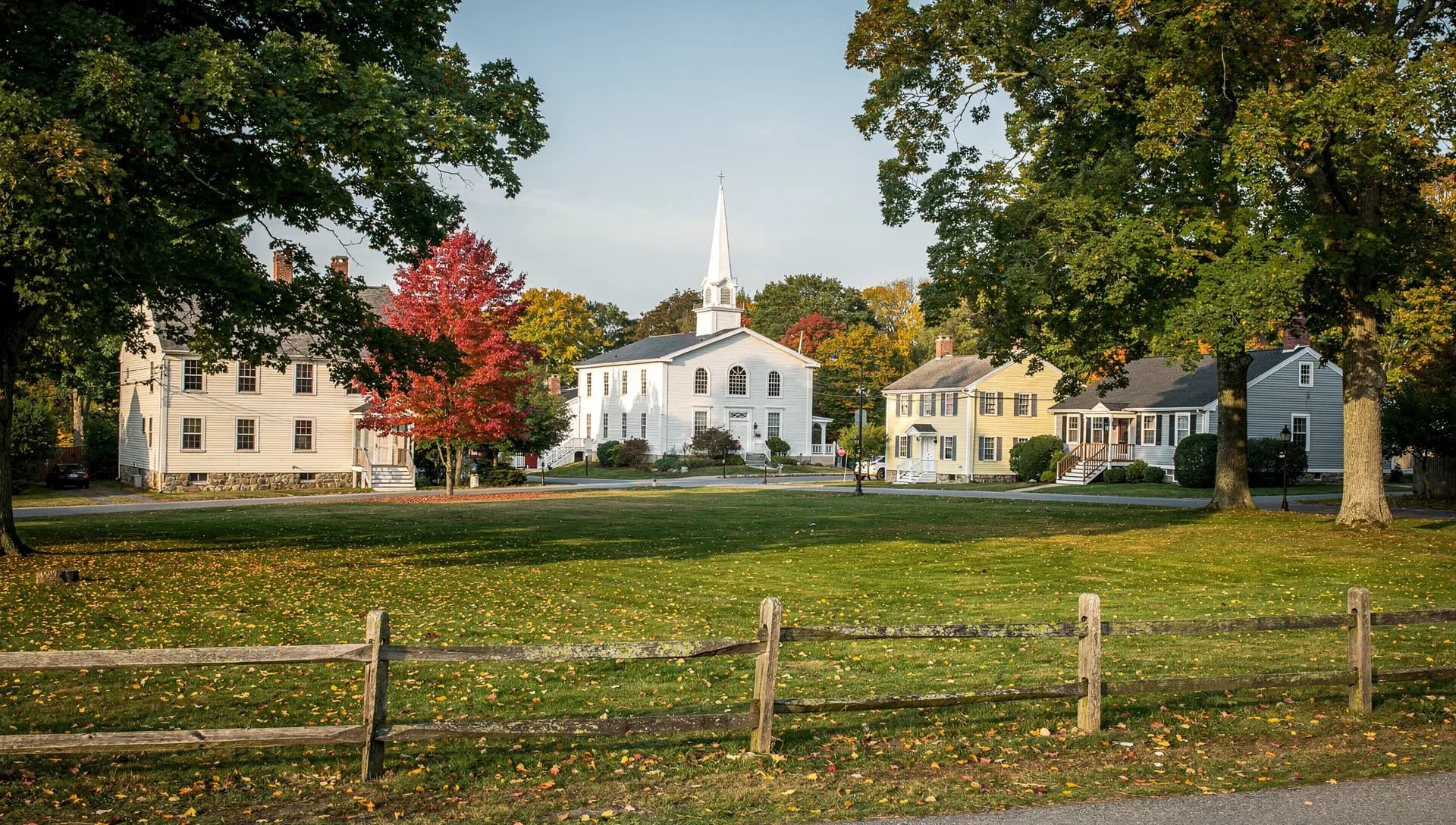 A town common lies behind a split-rail fence, with a white steepled church and old colonial houses under big maples.
