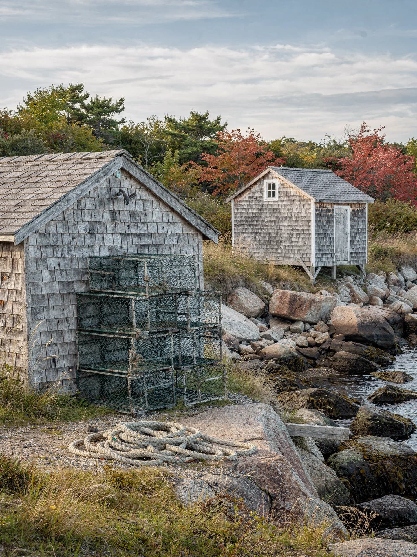 Two small weathered shingled fish houses stand above a rocky cove, wire lobster traps stacked against the nearer wall and a coil of rope on the ground.