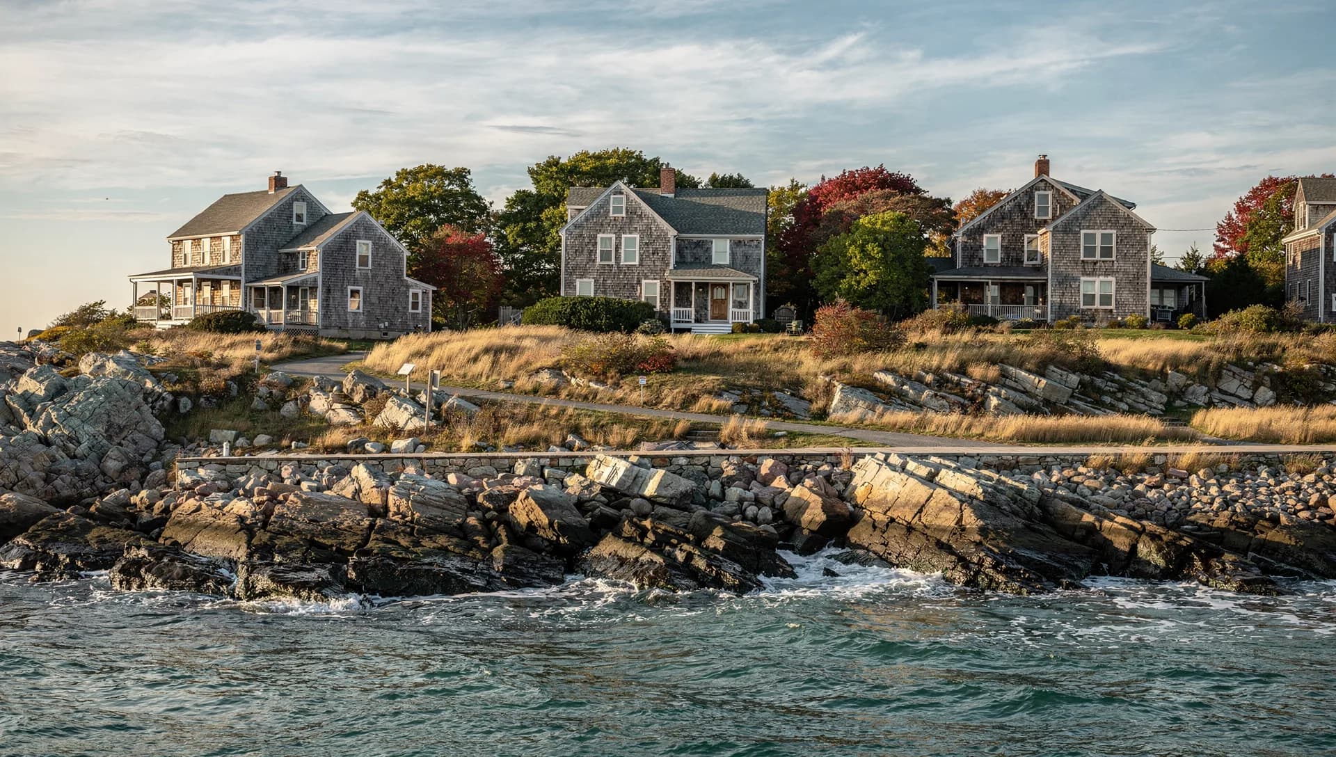 Shingled summer houses sit on a headland above a rocky shore, a footpath running along the top of the rocks.
