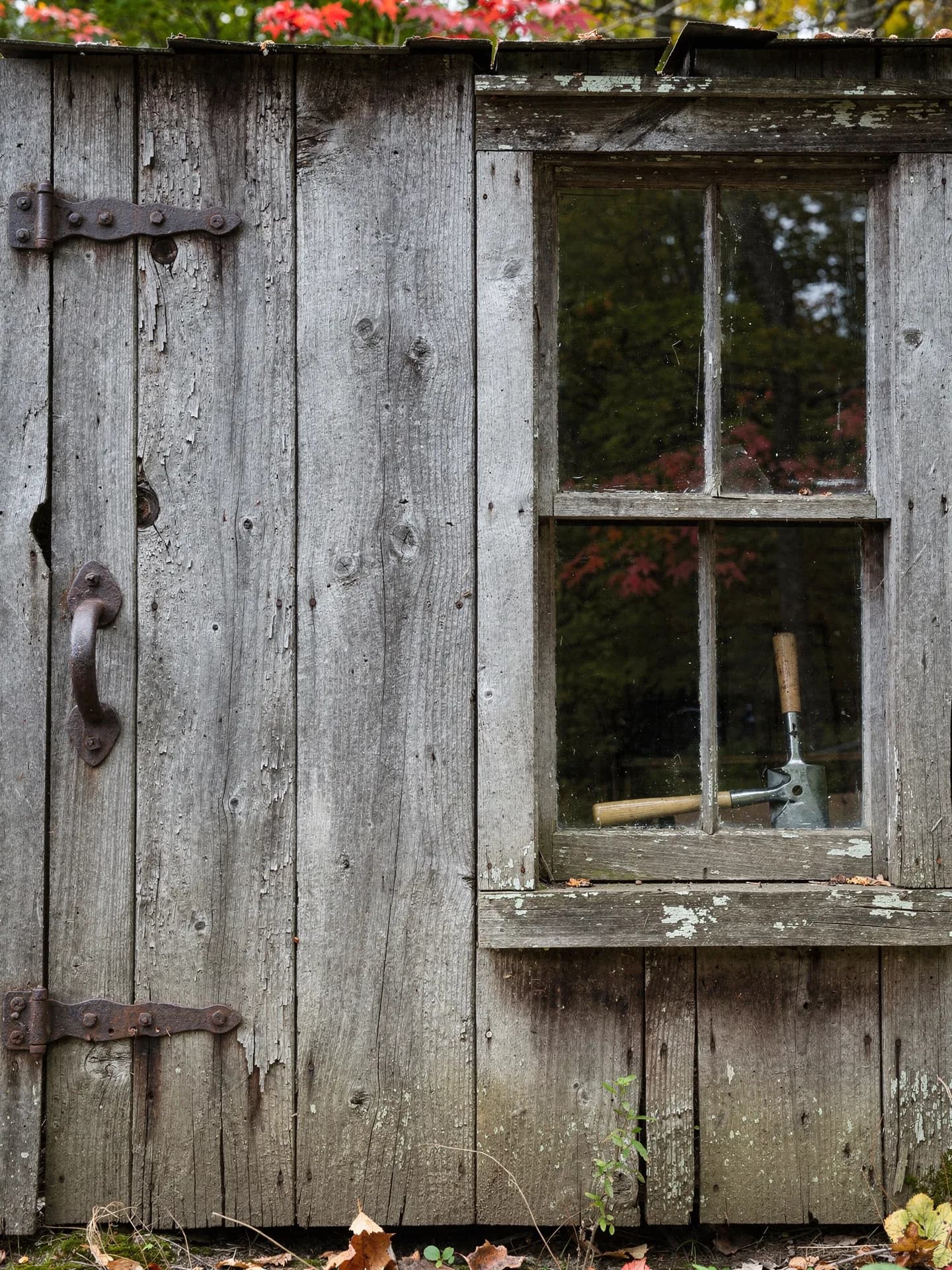 A weathered plank shed door with iron strap hinges and a forged handle stands beside a four-pane window, a hand tool resting on the sill inside.