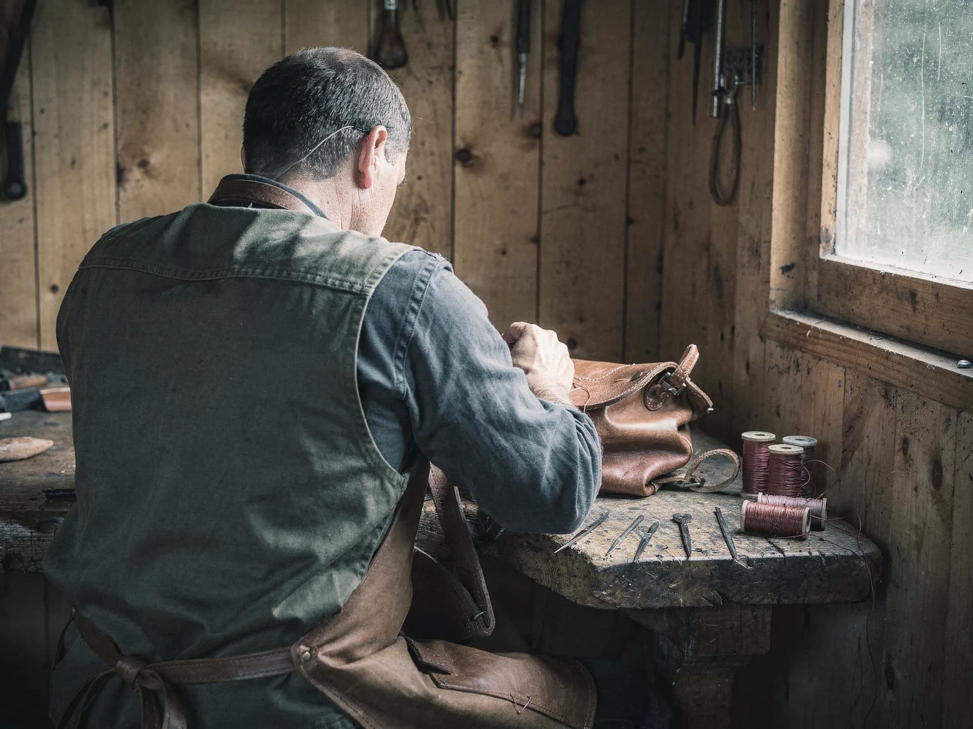 A leatherworker in a canvas vest and leather apron hand-stitches a brown leather bag at a bench by a window, awls and spools of thread laid out beside him.