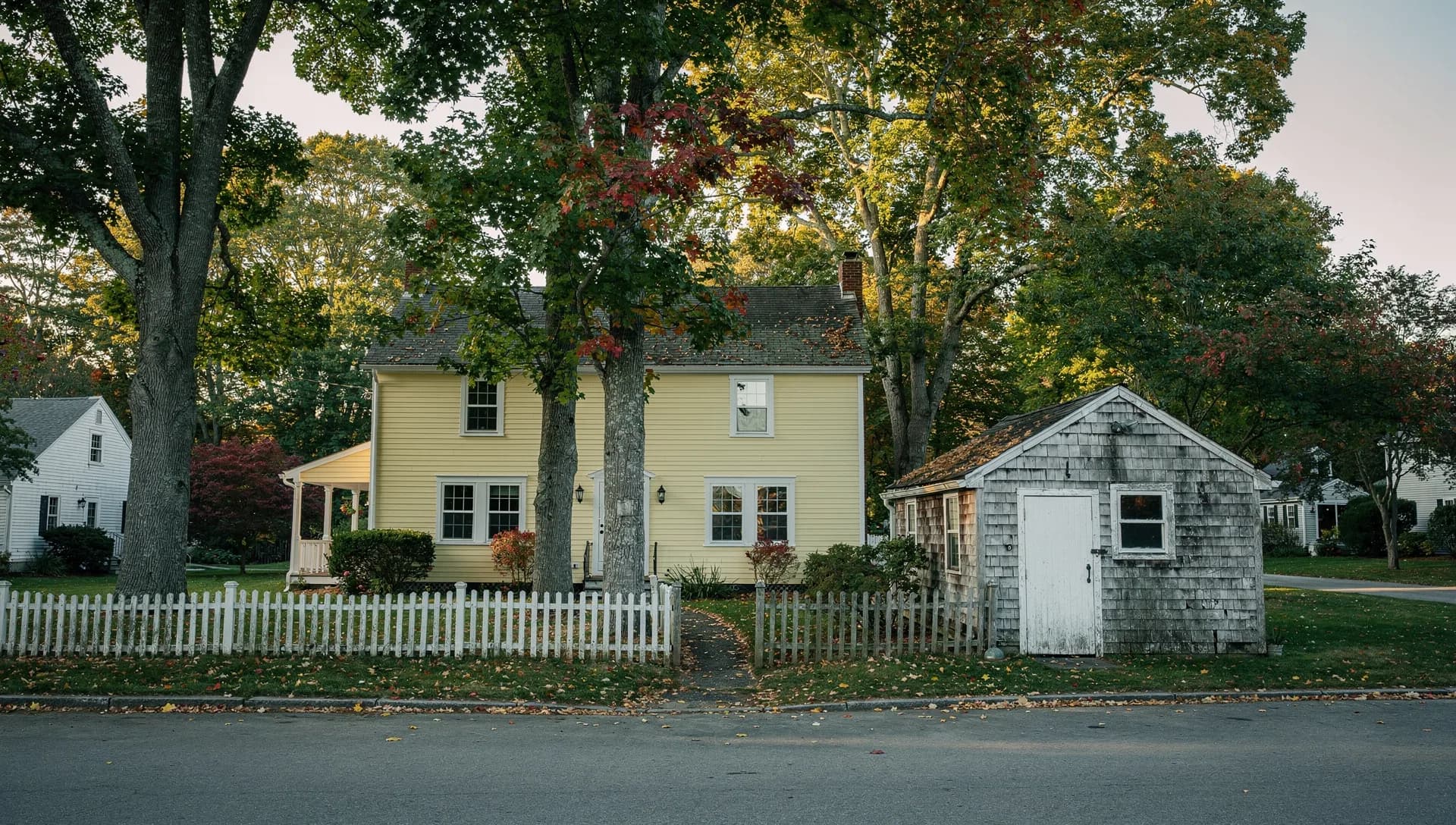 A pale yellow clapboard house and a small shingled workshop shed sit behind a white picket fence under tall maples.
