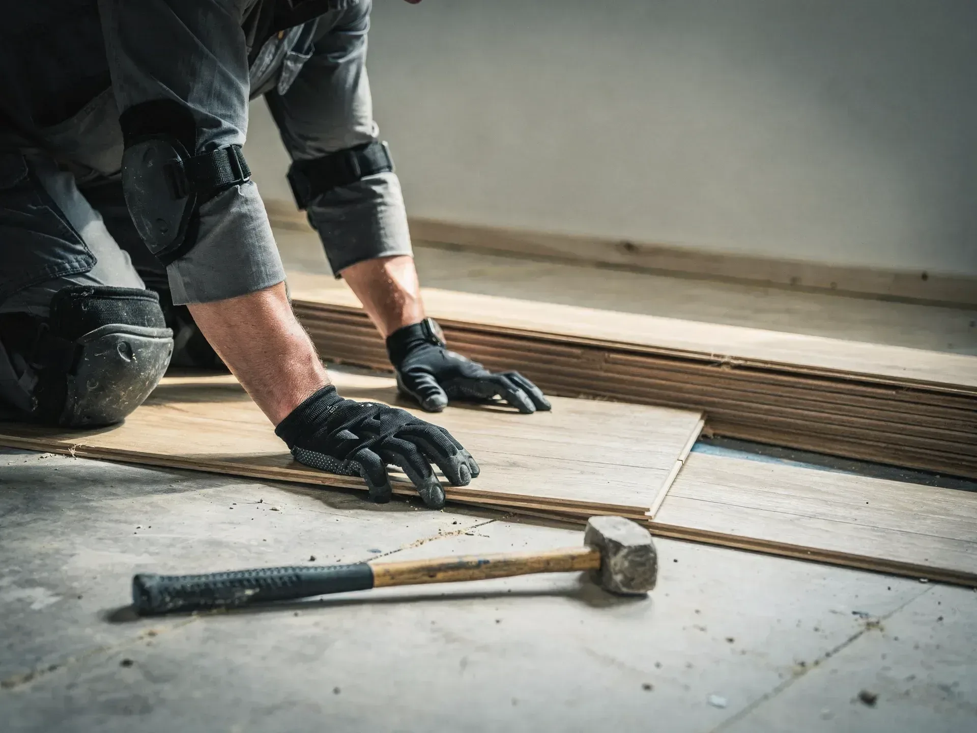 A flooring installer in knee pads and black work gloves kneels to press a pale oak plank into place beside a stack of planks, a mallet lying on the subfloor.