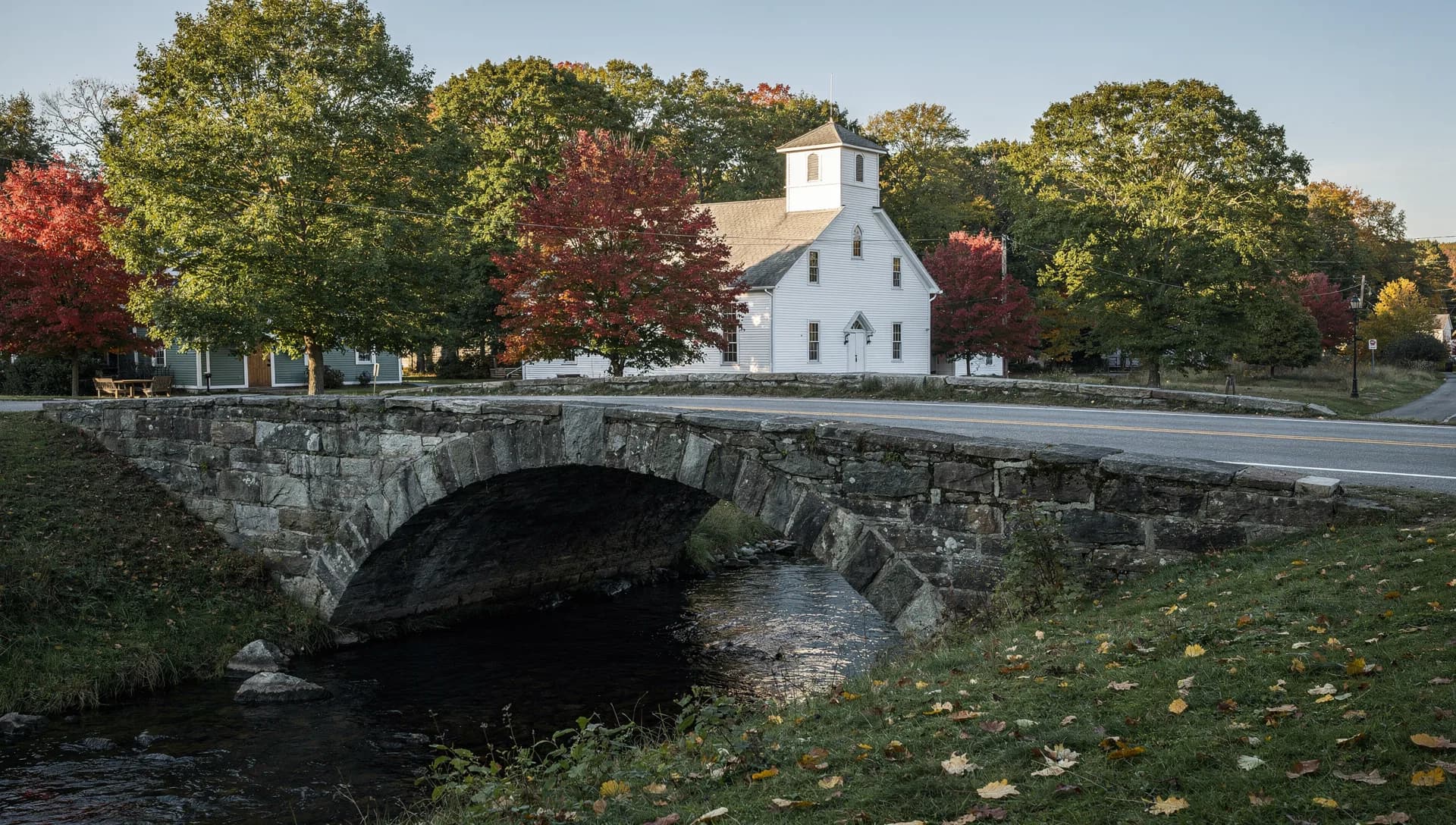A fieldstone arch bridge crosses a brook below a white church with a square tower, red maples around it.