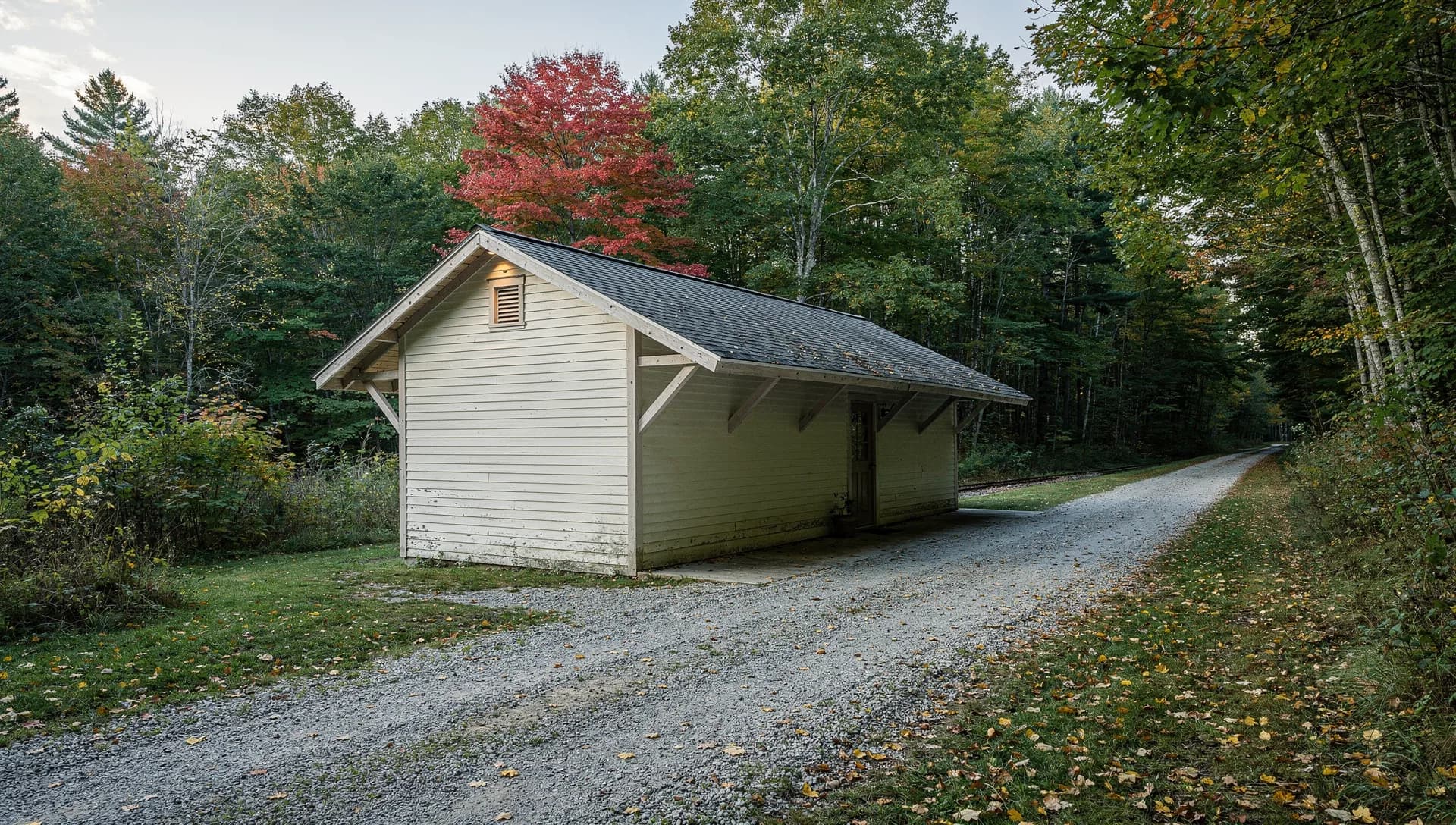 A small cream clapboard depot with a wide bracketed roof sits beside a gravel rail trail running into the woods.