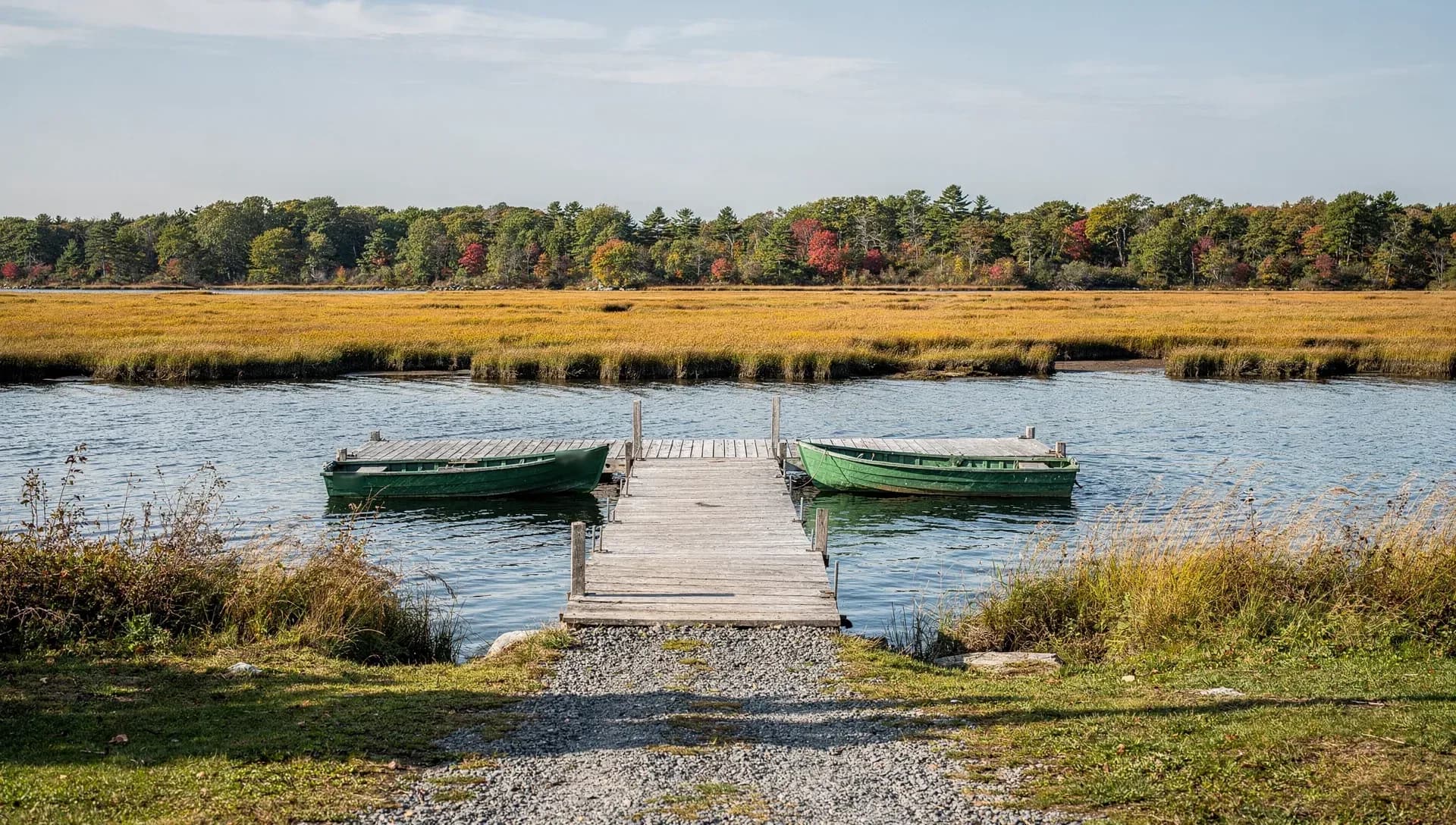 Two green skiffs are tied to a wooden float at a gravel landing, golden salt marsh running out to a far treeline.