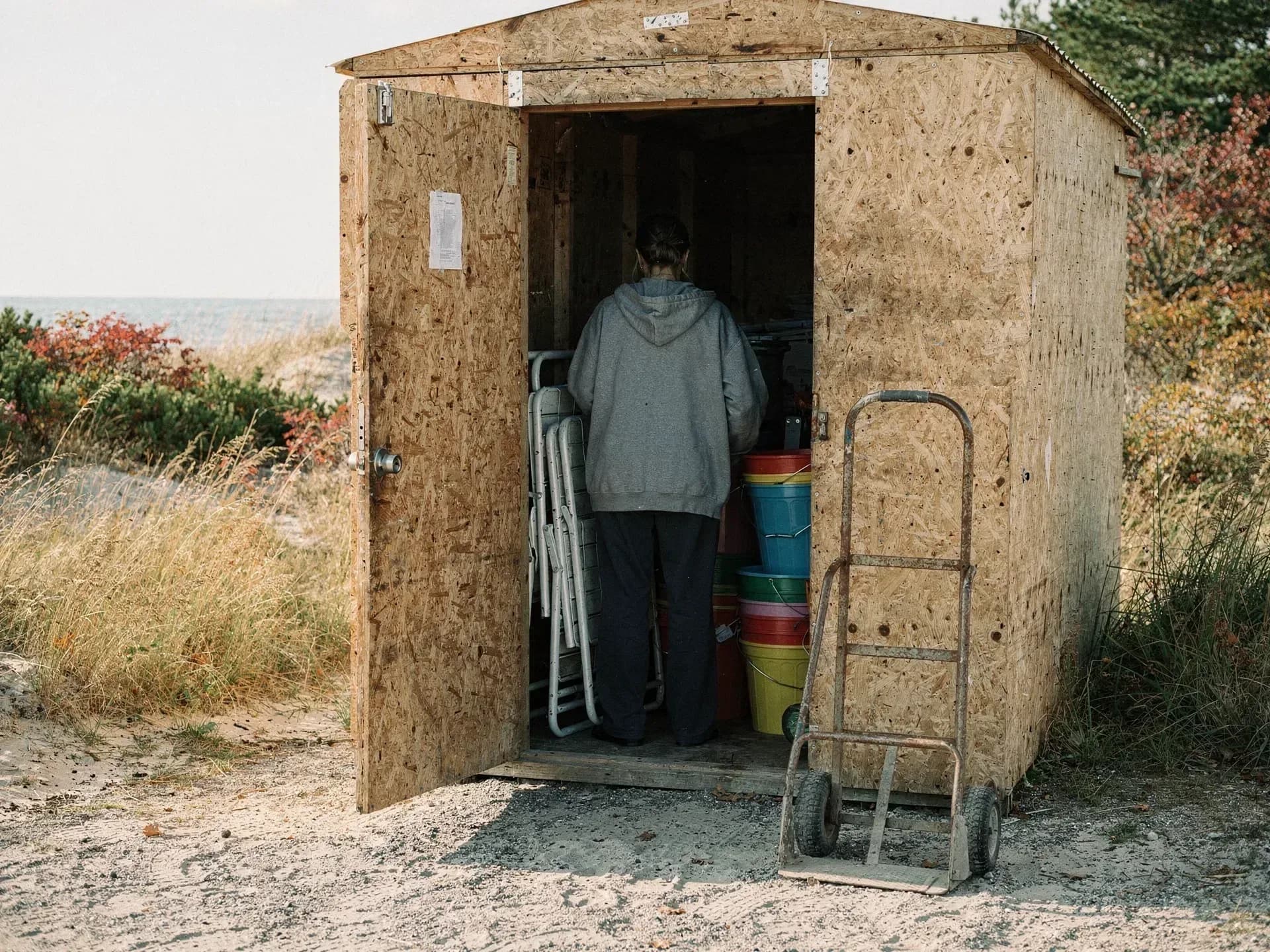 A woman in a grey hooded sweatshirt stacks folding chairs and bright plastic pails inside a plywood shed at the back of a beach, a hand truck leaning on the wall.