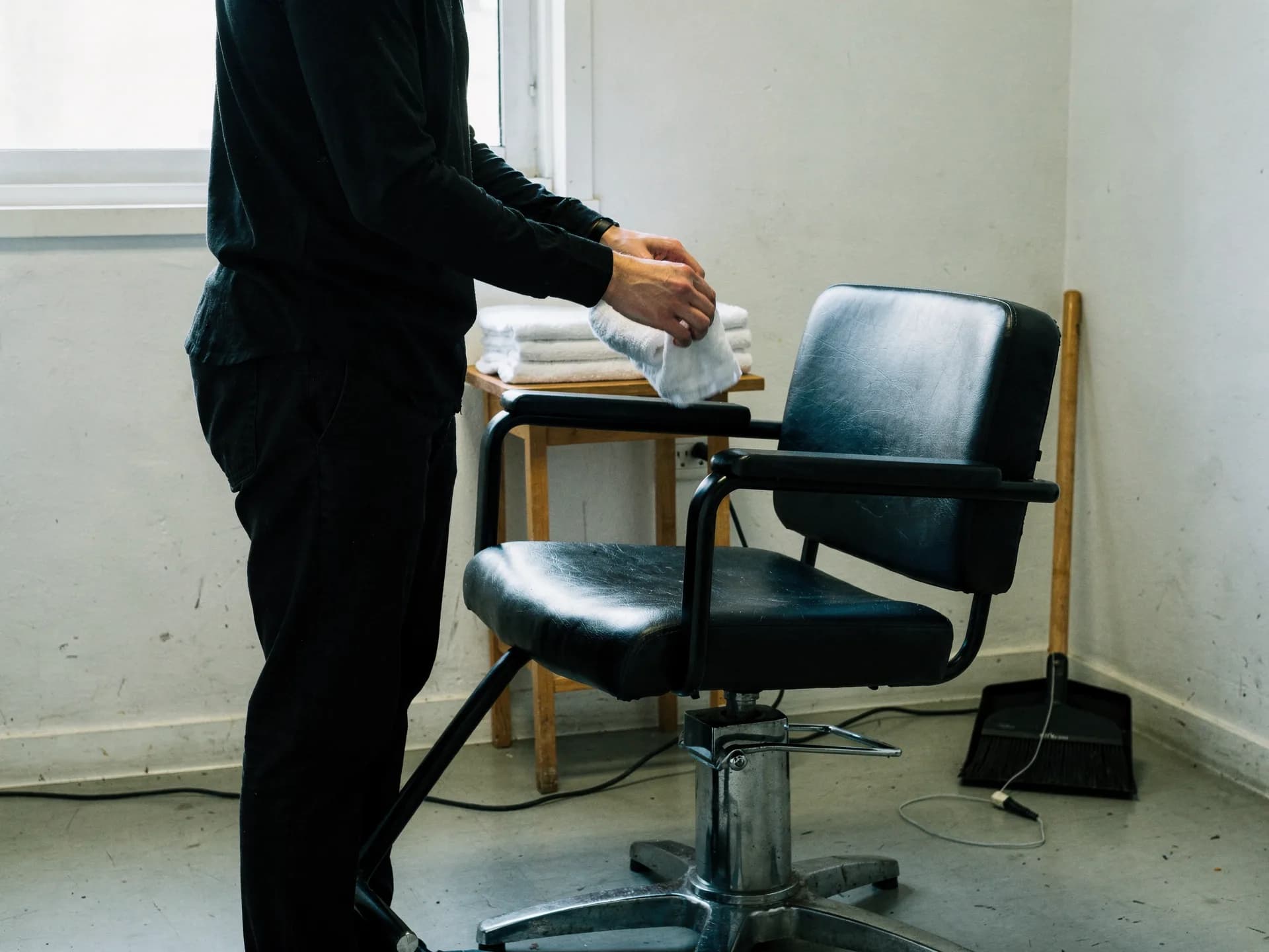 A stylist dressed in black folds a white towel onto a stack beside an empty black salon chair in a plain bright room, a broom and dustpan in the corner.