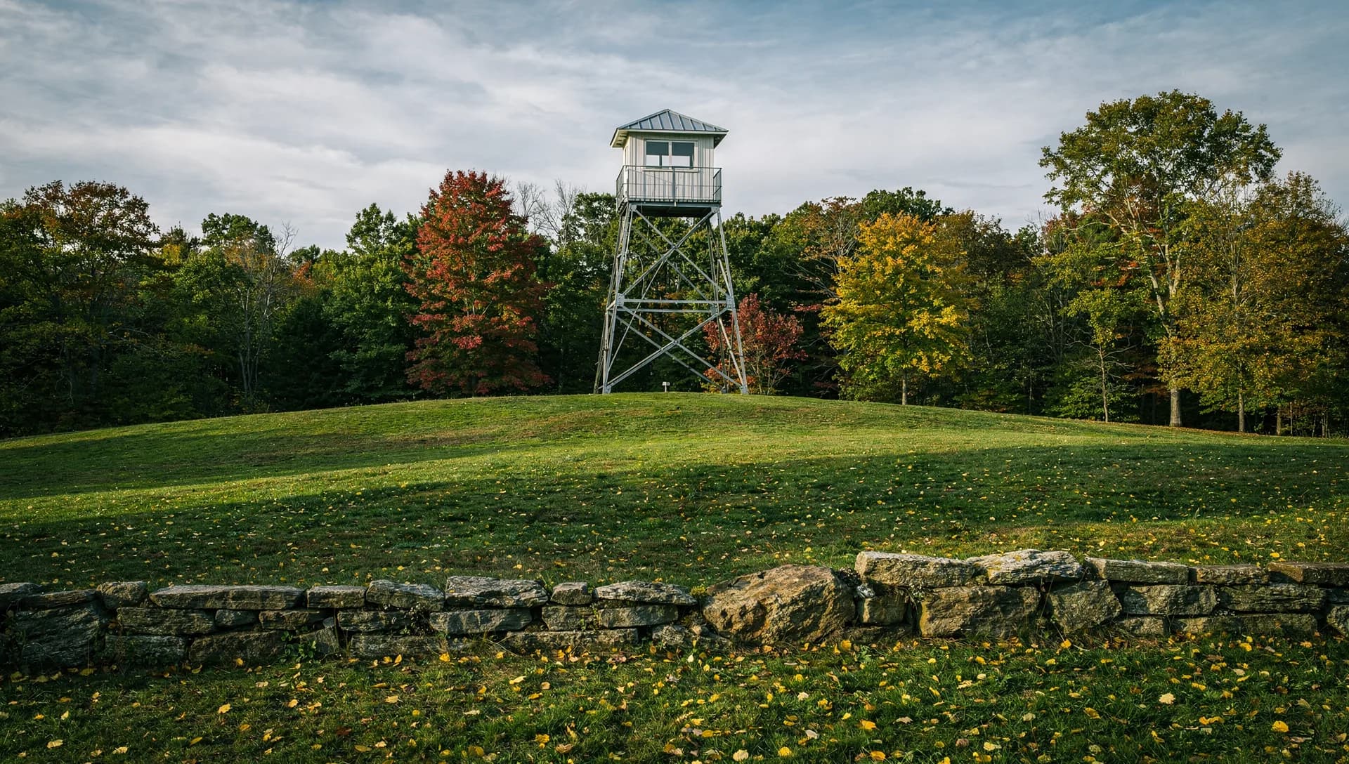 A steel lookout tower with a cabin on top stands on a mown hill behind a fieldstone wall, the woods around it turning.