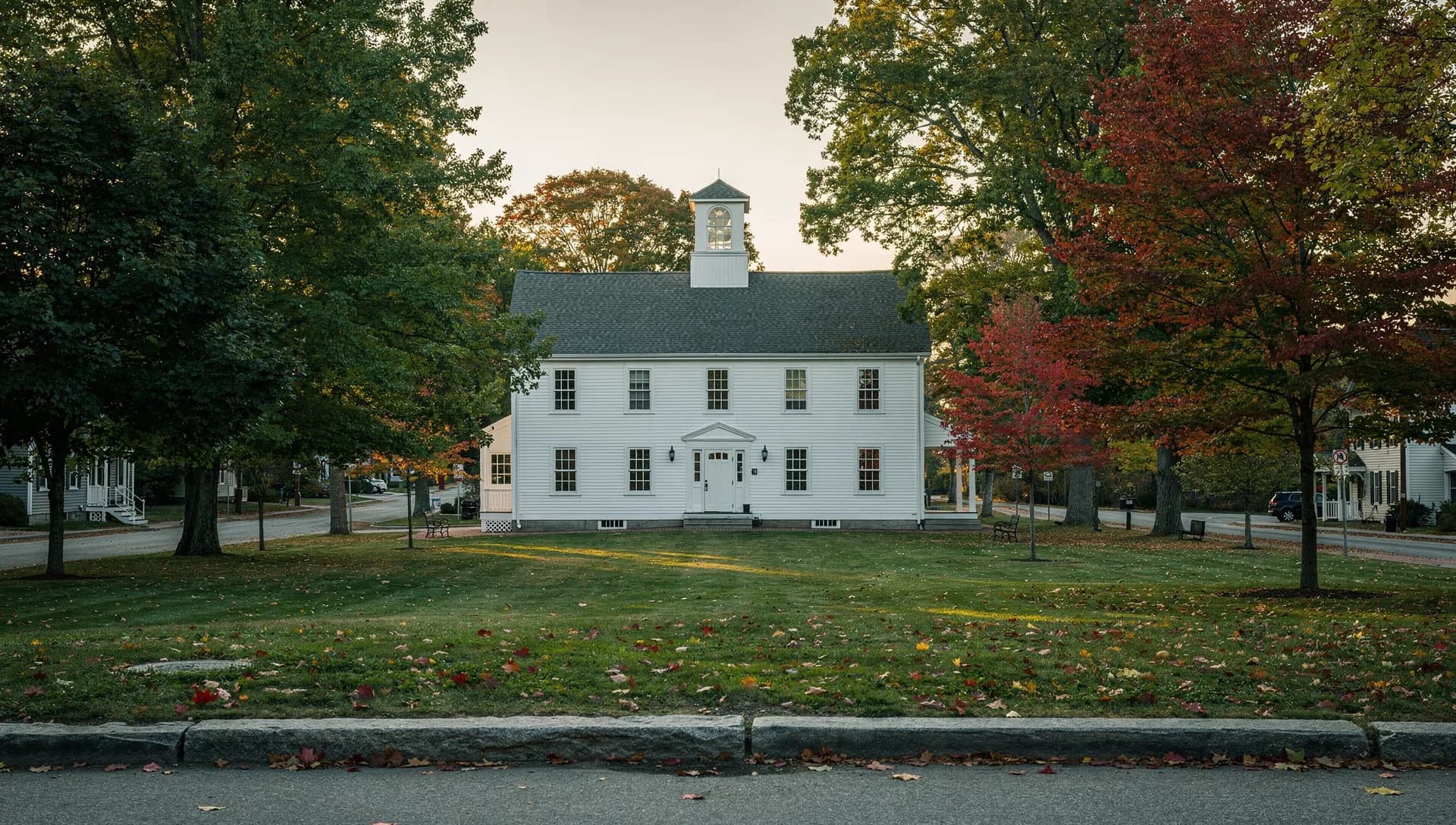 A white colonial town hall with a cupola stands on a green under maples, a granite curb along the street in front.