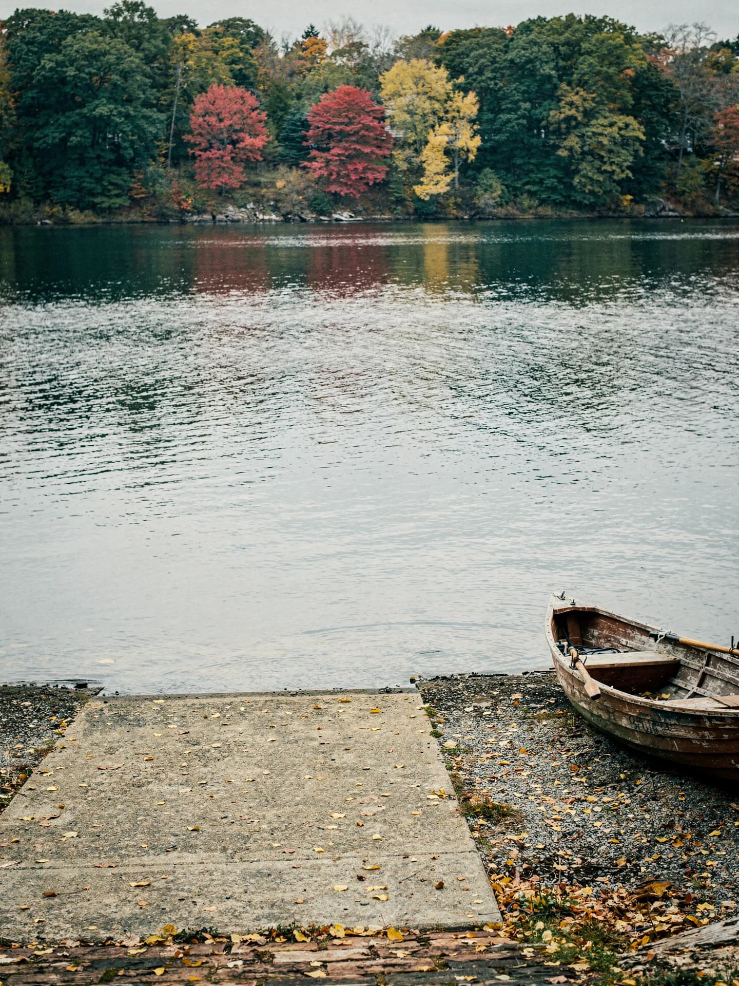 A concrete boat ramp slopes down to a wide river, a wooden rowboat pulled up on the gravel beside it and autumn trees on the far bank.