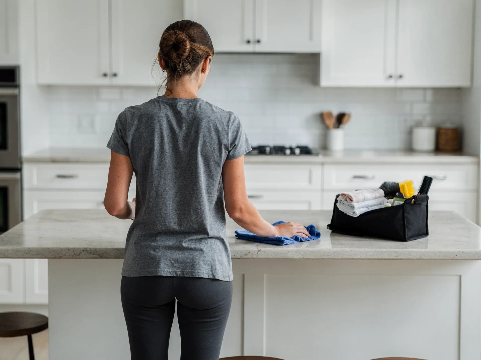 A house cleaner in a grey tee, her hair in a bun, wipes a stone kitchen island with a blue cloth, a caddy of cloths and brushes beside her.