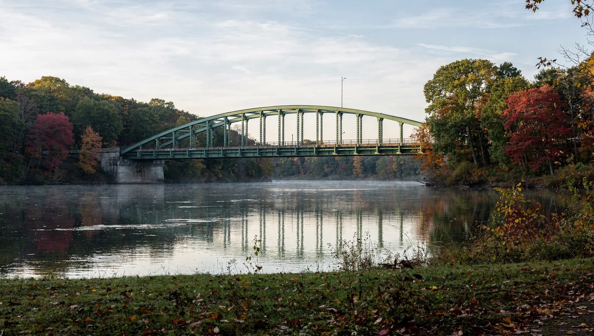 A green steel arch bridge spans a broad calm river and its own reflection, the wooded banks starting to turn.