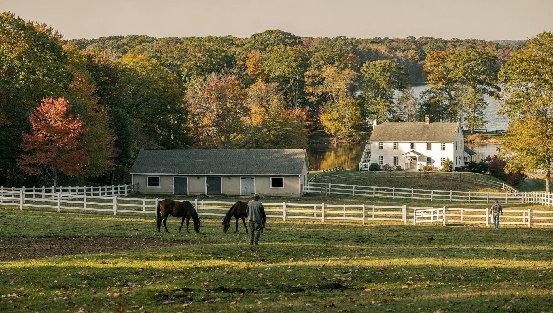 White fences divide a horse pasture beside a stable and a white farmhouse, a river beyond and two people walking in the distance.
