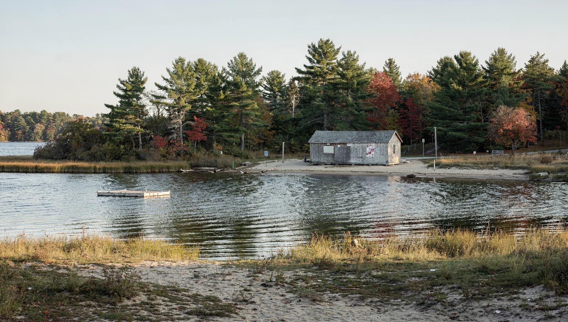 A small lake with a sandy beach, a weathered bathhouse and a swim float sits under white pines along the shore.