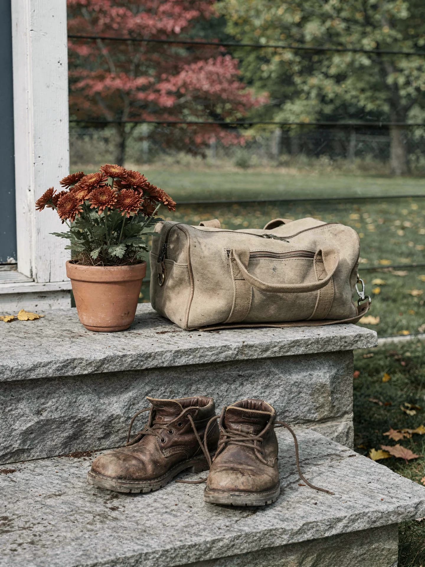 Worn leather work boots sit on a granite step below a canvas tool bag and a terracotta pot of rust-orange mums.