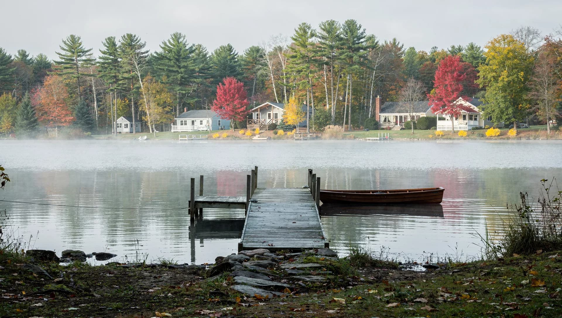 A wooden dock and a moored rowboat sit on a misty pond, cottages among white pines on the far shore.