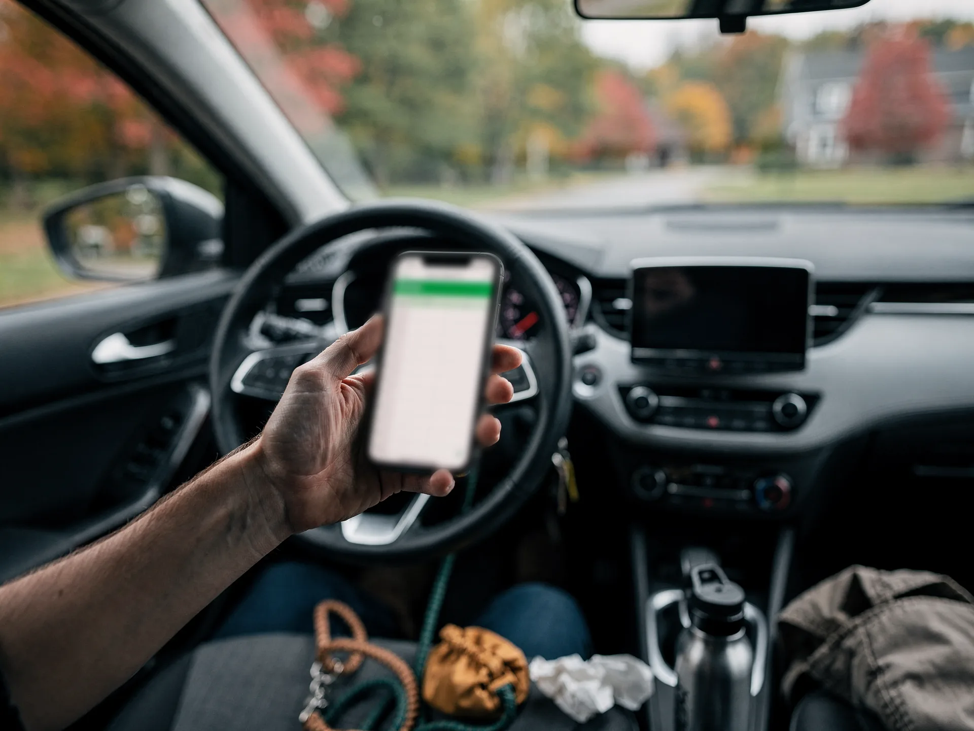 From the driver's seat of a car, a hand holds up a phone showing a spreadsheet with a green header, a coiled leash and a canvas pouch on the seat and autumn trees through the windshield.