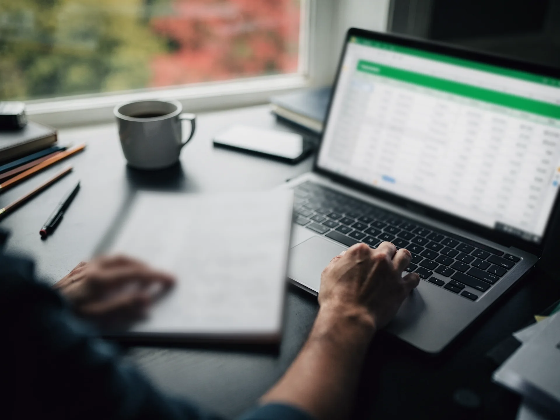 Hands at a laptop showing a spreadsheet with a green header bar, one resting on a spiral notebook of notes, a mug and pencils by a window.