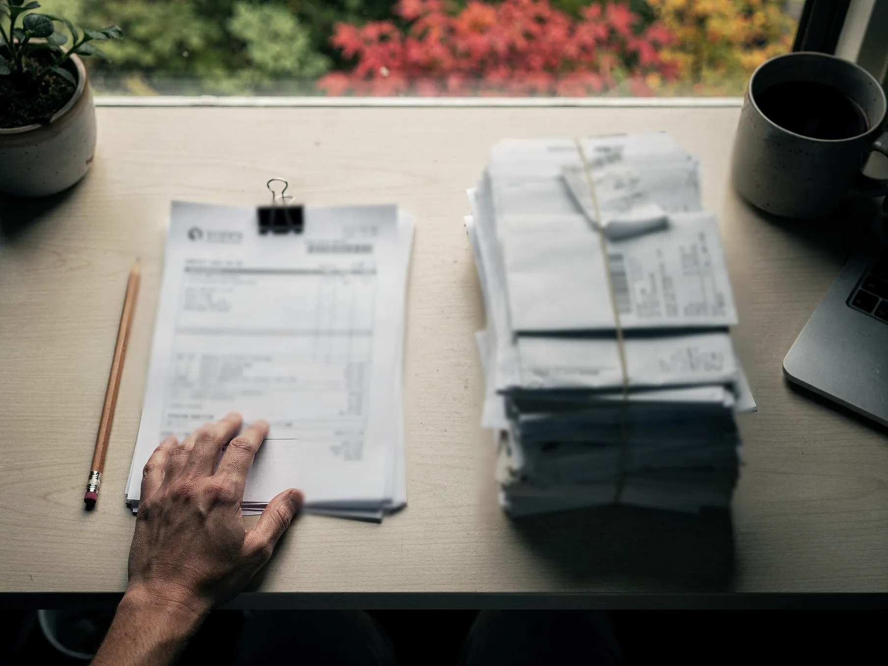 Overhead on a desk by a window, a hand rests on a binder-clipped stack of printed statements beside a thick bundle of receipts and envelopes tied with string, a pencil and a mug nearby.
