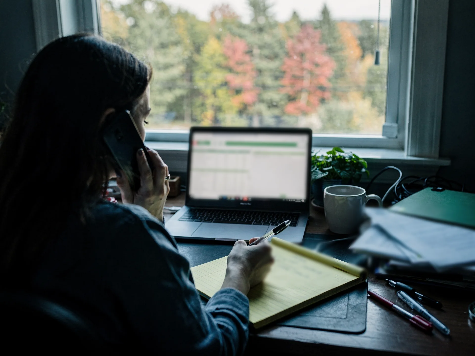 A woman with long dark hair, turned away, talks on the phone at a window desk, pen over a yellow legal pad, a laptop showing a green-header spreadsheet, a mug and a pile of papers beside her.