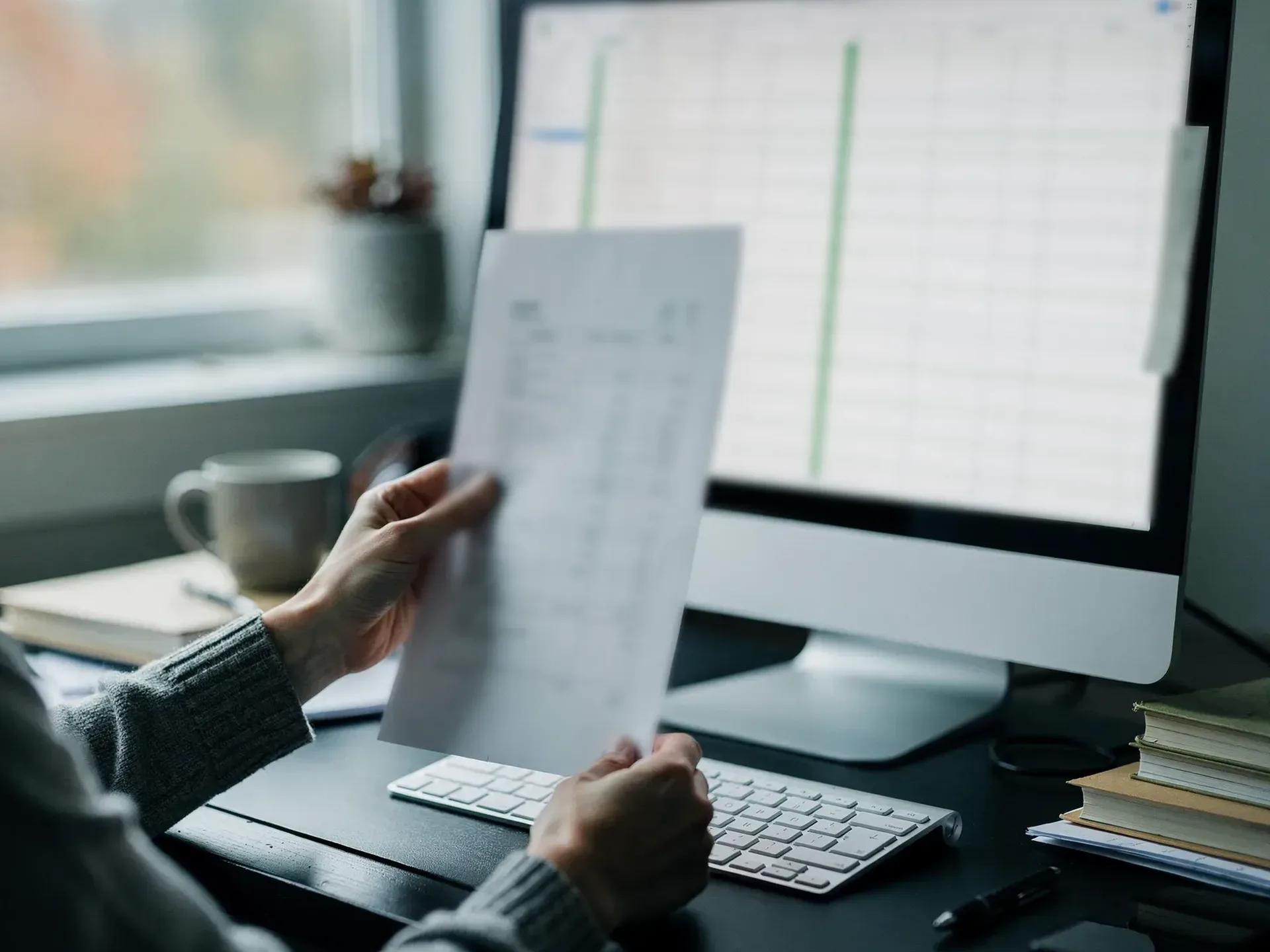Hands in grey knit sleeves hold a printed statement up beside a monitor showing a ledger with green columns, a mug and books on the desk by a window.