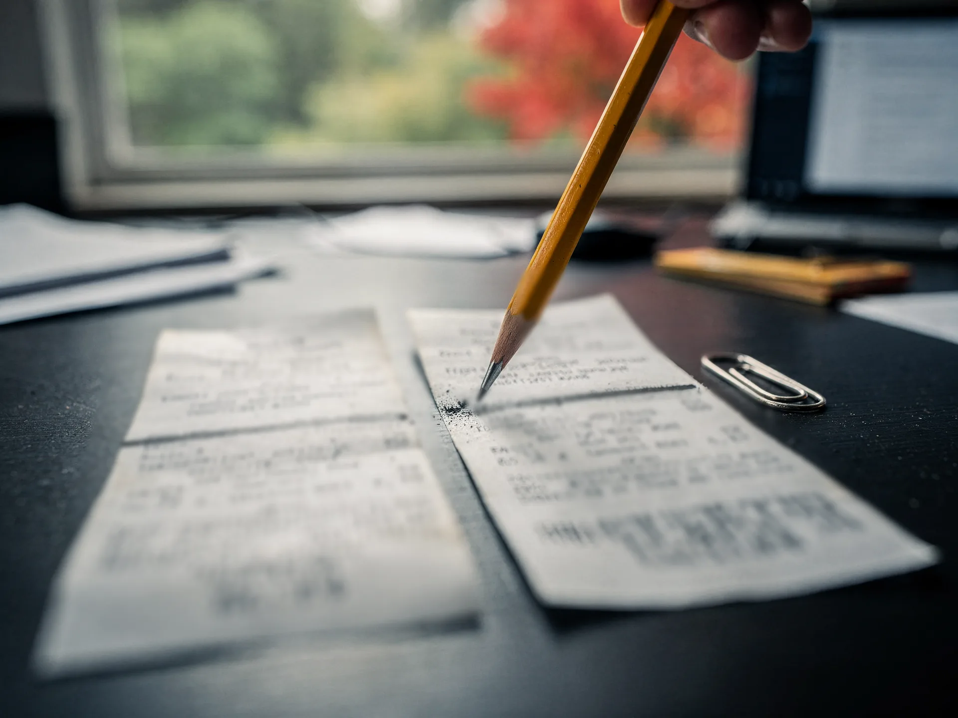 A pencil draws a line across one of two faded paper receipts laid side by side on a dark desk, a paper clip beside them.
