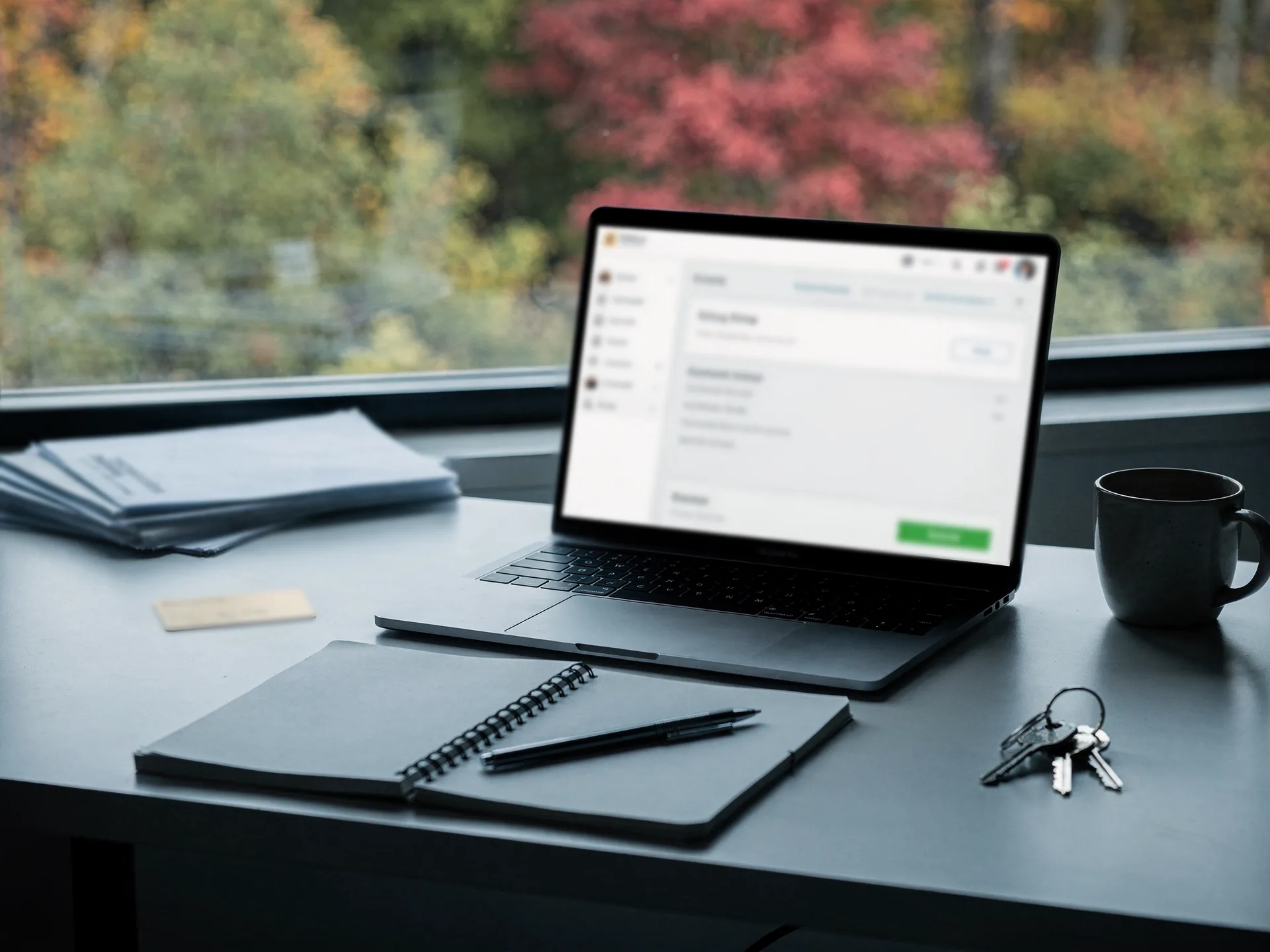 An open laptop showing a settings screen with a green button sits on a dark counter by a window, beside a spiral notebook and pen, a stack of envelopes, a card, keys and a mug.